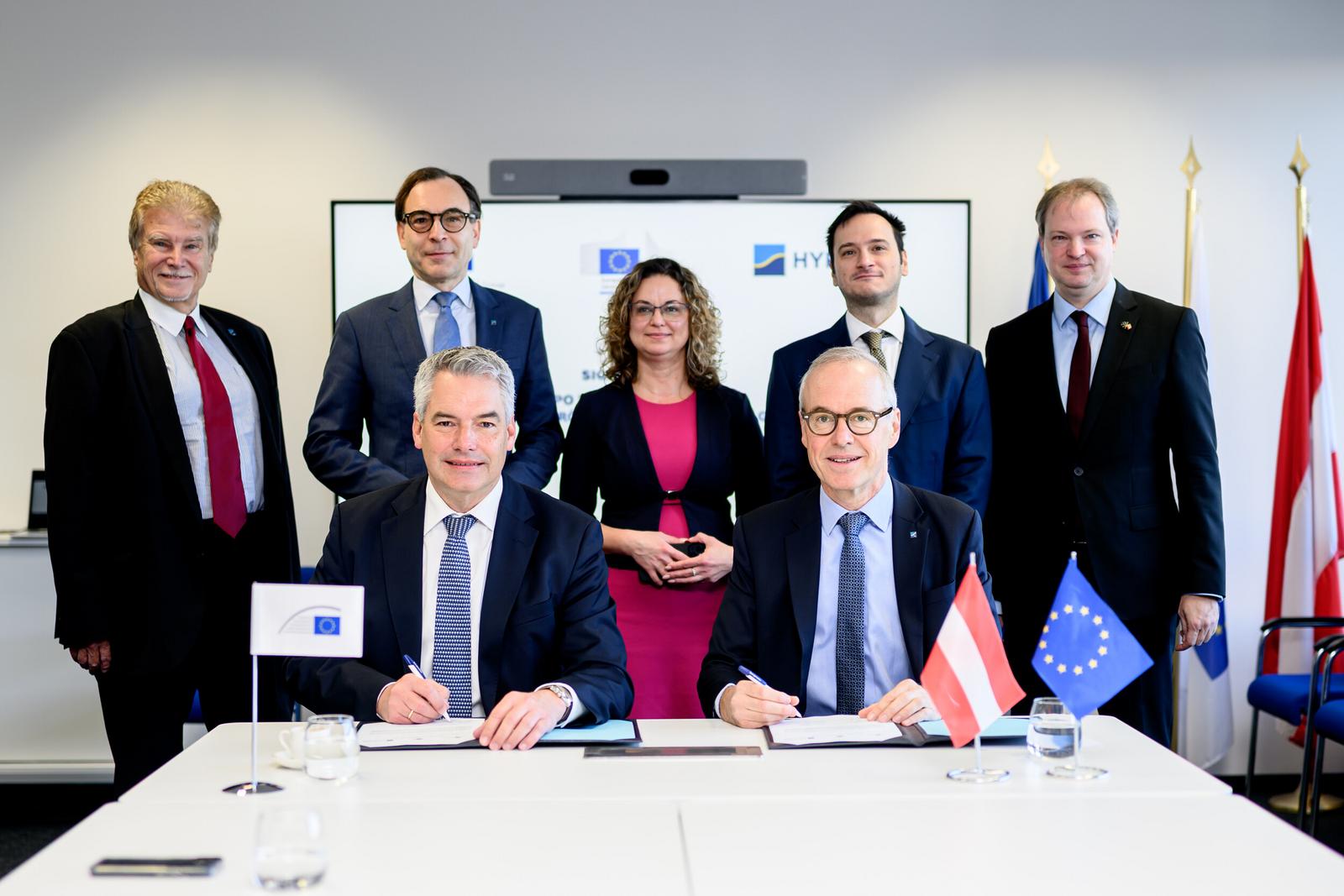 Zwei Männer in Anzug und Krawatte sitzen an einem Tisch, dahinter stehen vier weitere Männer in Anzug und Krawatte und eine Frau.