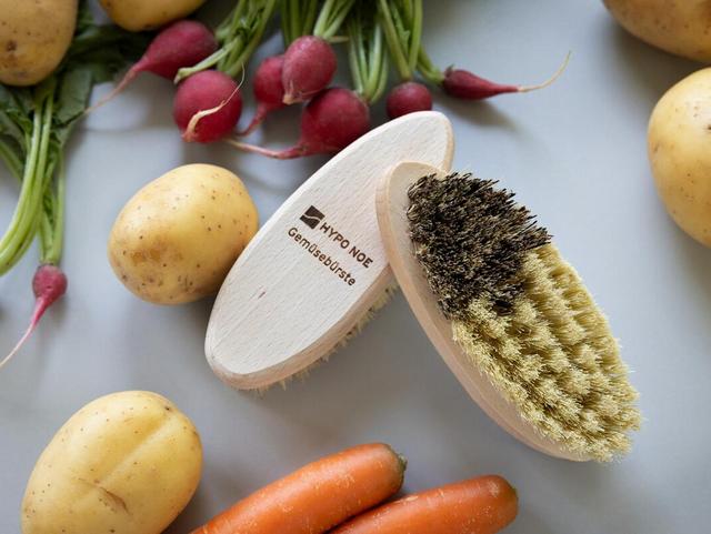 zwei Bürsten aus Holz mit groben Borsten liegen zwischen Kartoffeln, Karotten und Radieschen