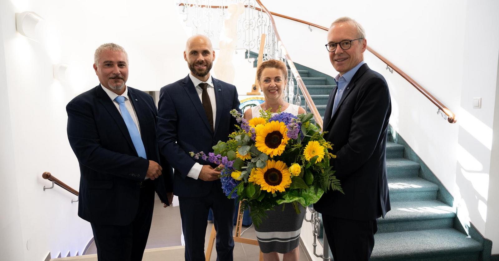 Drei Männer und eine Frau in Businesskleidung - die Frau hält einen großen Blumenstrauß.