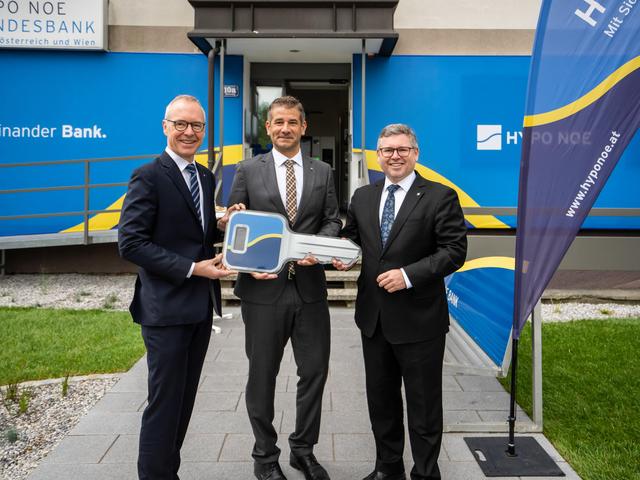 Drei Männer mittleren Alters in Anzügen stehen vor einem Gebäude, dessen Fassade mit dem blau-gelben Logo der HYPO NOE versehen ist.