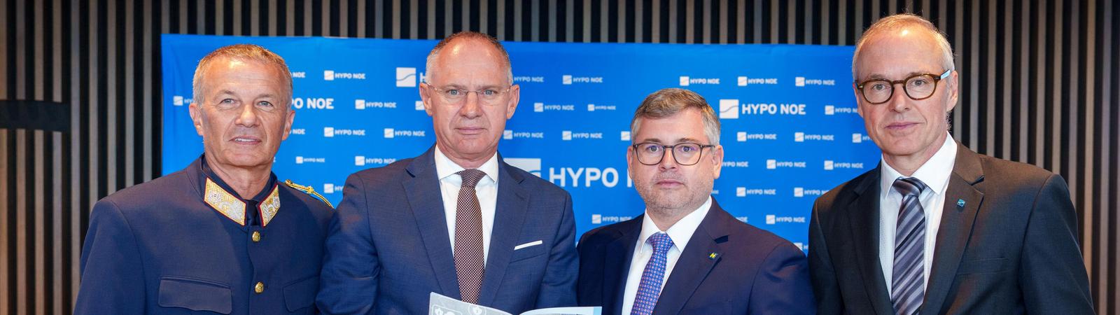 Vier Herren im Anzug stehen vor einem Pult und halten eine Broschüre in Händen.