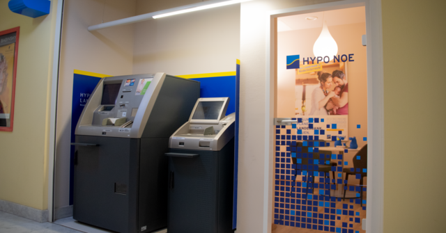 zwei große Geräte - ein Bankomat und ein Kontoauszugsdrucker - stehen in einer Wandnische neben einer Glastür