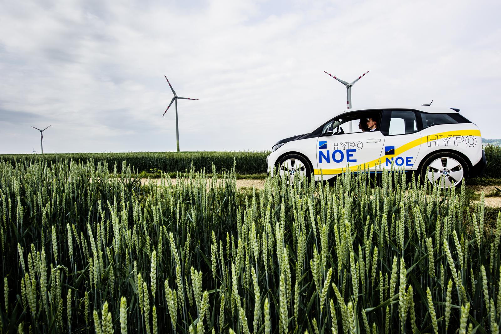 Ein Elektrofahrzeug aus dem Fuhrpark der HYPO NOE fährt durch grüne Wiesen - im Hintergrund Windräder
